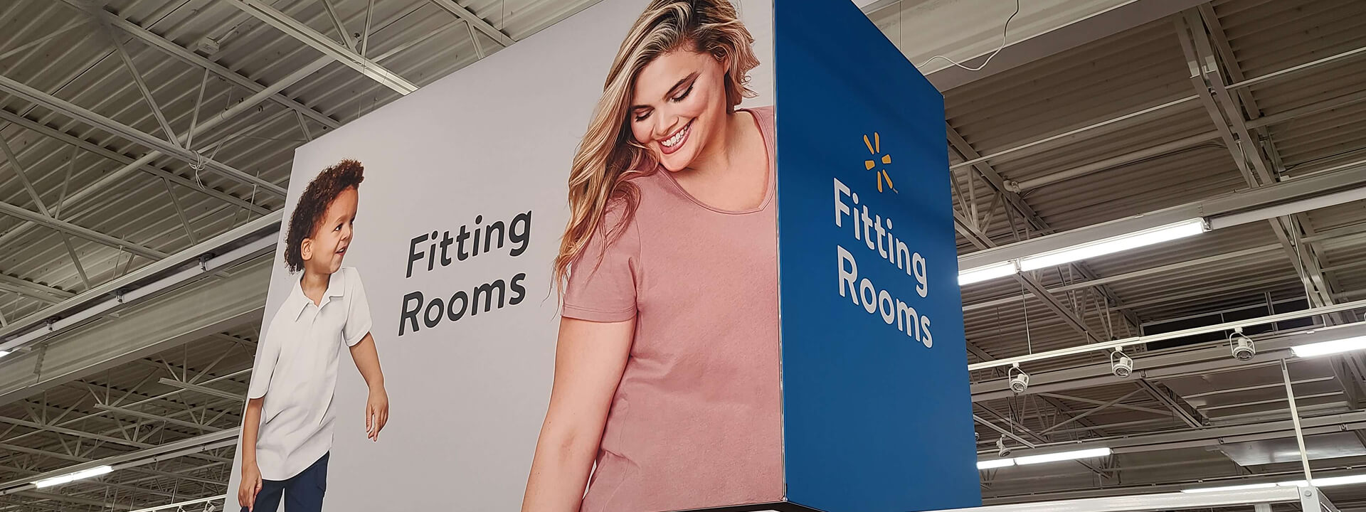A large hanging sign in a retail store displays ""Fitting Rooms"" with images of a smiling woman and child, and the Walmart logo on a blue panel.