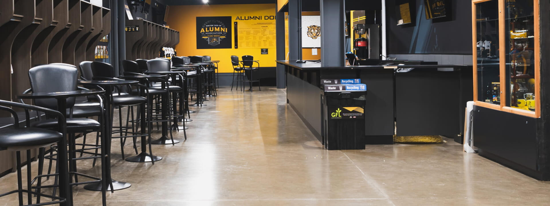A modern alumni clubhouse with black high-top tables and stools, dark wood shelving, a yellow wall with ""Alumni"" branding, and a service counter with recycling bins.