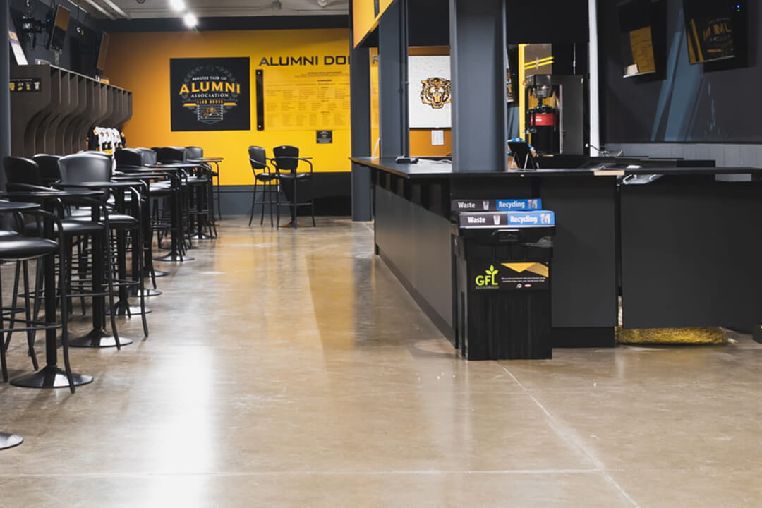 An empty modern lounge with rows of black high-top tables and stools, yellow walls featuring ""Alumni Association"" signage, and a service counter with recycling bins.