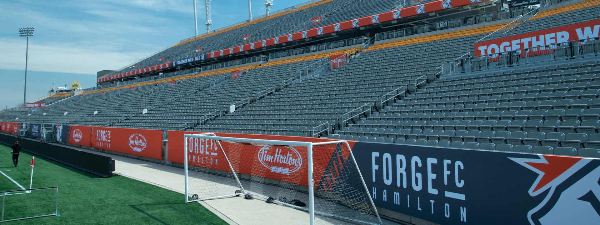 An empty soccer stadium features vast grey seating, a goal on a green field, and Forge FC and Tim Hortons Field banners.