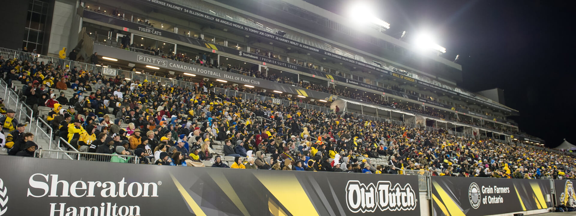 A brightly lit stadium at night is filled with a large crowd of fans, many wearing black and yellow, with prominent signs for the Canadian Football Hall of Fame Club and the Tiger-Cats visible in the stands.