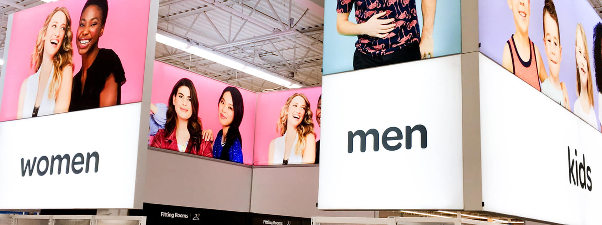 Overhead store signs display sections for ""women,"" ""men,"" and ""kids,"" each featuring corresponding images of smiling models.