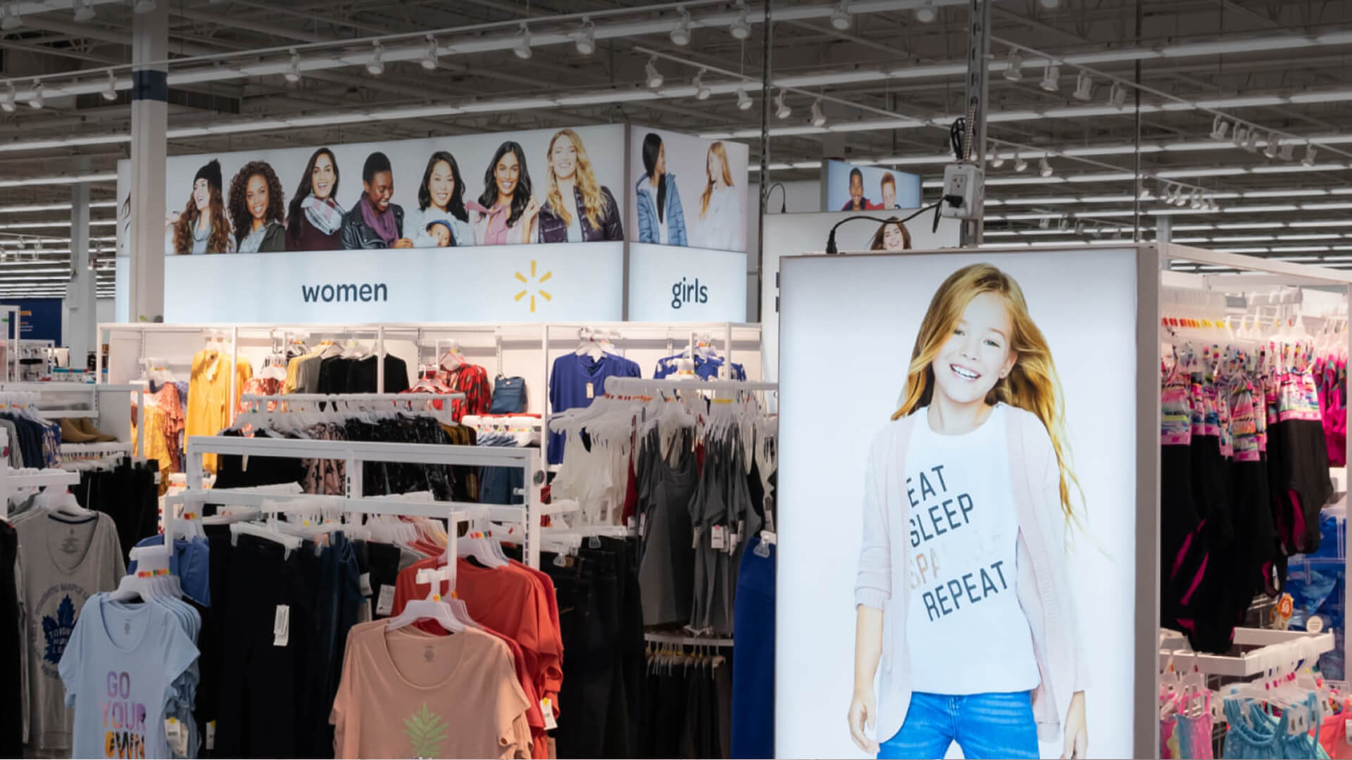 A brightly lit clothing department with ""women"" and ""girls"" signs, multiple clothing racks, and a large display featuring a smiling girl.
