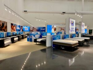 "A brightly lit Rooms To Go Outlet Sleep Center showroom featuring rows of mattresses, branded banners, and large screens displaying sleep-themed imagery."