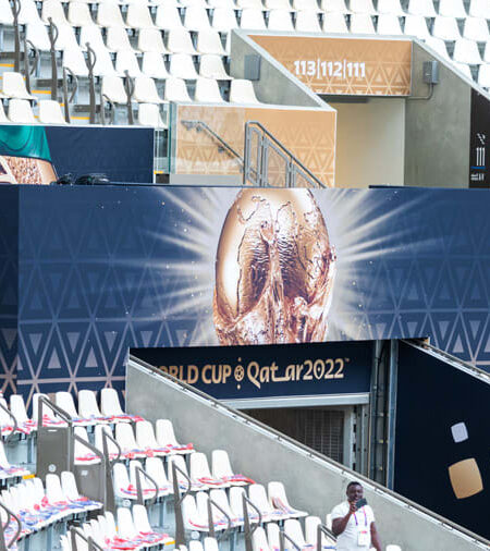 Empty stadium stands with a large banner displaying the FIFA World Cup trophy and 'Qatar 2022', overlooked by a camera crew.