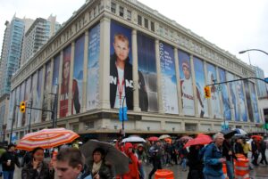 A large building featuring Vancouver 2010 Olympic athlete banners overlooks a rainy city street filled with pedestrians carrying umbrellas.