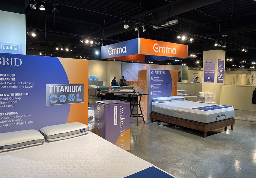 An Emma Sleep brand trade show booth displaying multiple mattresses, pillows, and product information.