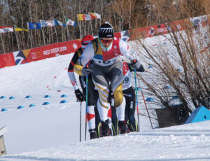"A cross-country skier in a multi-colored suit powers uphill on a groomed track, followed by other racers, with ""Red Deer 2019"" banners in the background."