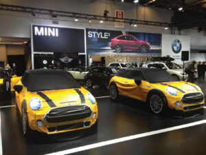 Two yellow Mini Cooper display models with black roofs are prominently featured at a car exhibition.