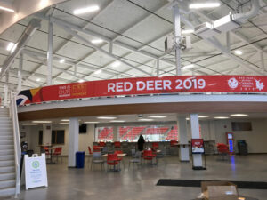 "An interior view of an arena concourse features a large ""RED DEER 2019"" banner, a lounge area with tables and chairs, and a partial view of red stadium seats."