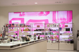 A modern beauty product display featuring shelves of perfumes and cosmetics against a vibrant pink-lit wall with abstract bottle designs.