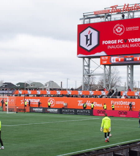 An inaugural soccer match between Forge FC and York9 FC is displayed on a large scoreboard at Tim Hortons Field, with players on the pitch and spectators in the stands.