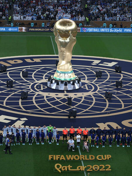 The golden FIFA World Cup trophy is displayed on a ceremonial pitch, with football teams and spectators in the stadium at the Qatar 2022 final.