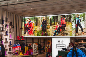 "Interior of a Hunter store displaying colorful wellington boots and rain jackets, with a large advertising mural of models in a forest setting."
