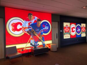 "A large illuminated display features two hockey players (Calgary Flames and Winnipeg Jets) and their logos, alongside smaller panels for the 2019 Heritage Classic."