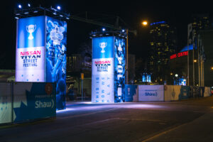 "Tall, blue-lit banners for the Nissan Titan Street Festival and Grey Cup Festival line a dark city street at night, with the Rogers Centre visible in the background."