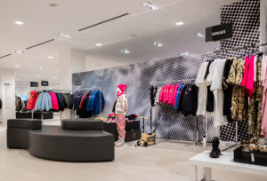 "A modern luxury clothing store interior features racks of colorful apparel, a mannequin in a pink hat, a black and white polka dot wall, and a Versace sign."