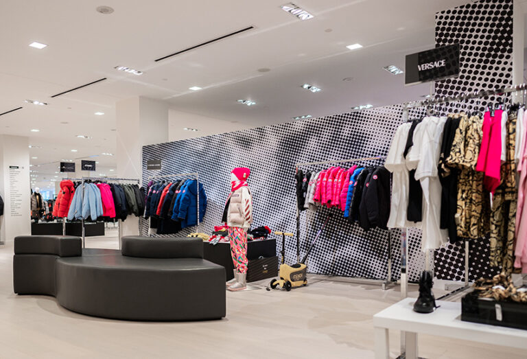 "A modern luxury clothing store interior features racks of colorful apparel, a mannequin in a pink hat, a black and white polka dot wall, and a Versace sign."