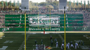 "A football game in a green stadium packed with fans, featuring players on the field and ""Pil Country"" banners."