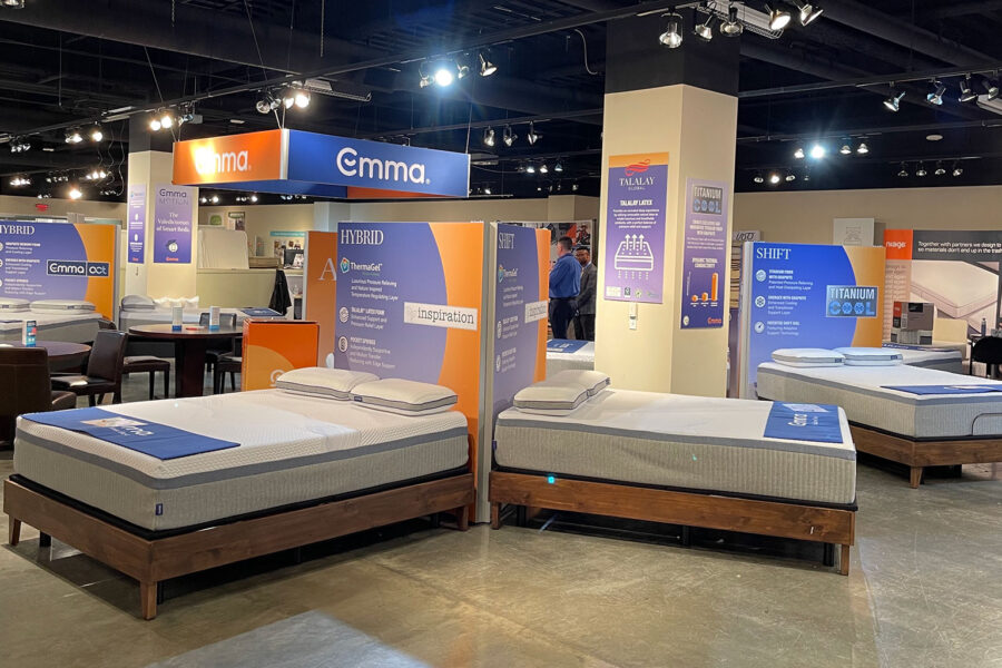 A brightly lit mattress showroom features multiple beds on wooden frames alongside promotional banners for brands like Emma, Hybrid, and Titanium Cool.