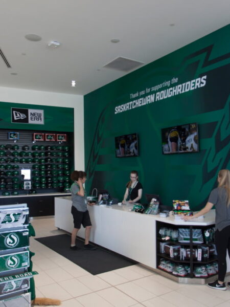A bright Saskatchewan Roughriders team store interior displays green and white merchandise, with staff assisting customers at the counter.