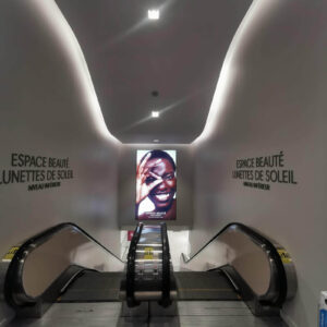 "Two escalators descend towards a large digital advertisement featuring a smiling woman making an ""OK"" gesture, flanked by French signs for ""Espace Beauté"" (Beauty Space) and ""Lunettes de Soleil"" (Sunglasses) pointing to the lower level."