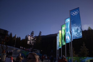 Blue and green Olympic flags fly over an outdoor event with people and a dark mountain under a clear blue sky.