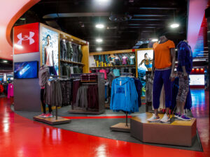"A vibrant Under Armour retail store interior showcases athletic apparel on mannequins and racks, with digital displays and a bright red floor."