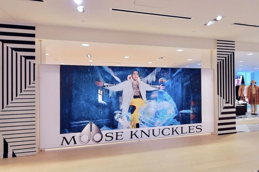 A retail store display features a large ""MOOSE KNUCKLES"" banner with a model in a white jacket and yellow pants amidst ice, framed by black and white striped columns, and clothing racks in the background.