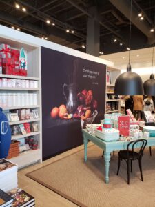 "A retail store interior shows a large still-life mural behind a display table with serveware on sale, next to shelves of home decor and packaged goods."