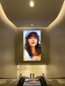 "An escalator descends into a brightly lit, modern space featuring a large, illuminated advertisement for ""Espace Beauté"" with a woman's face on the wall ahead."
