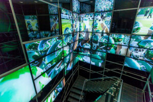 "A multi-level room features a large video wall displaying green-uniformed football players and game scenes, with a modern staircase descending in the foreground."