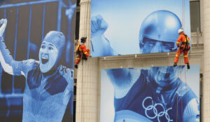 "Two workers in orange safety gear rappel down a building adorned with large blue-toned banners of celebrating athletes, including one with Olympic rings."