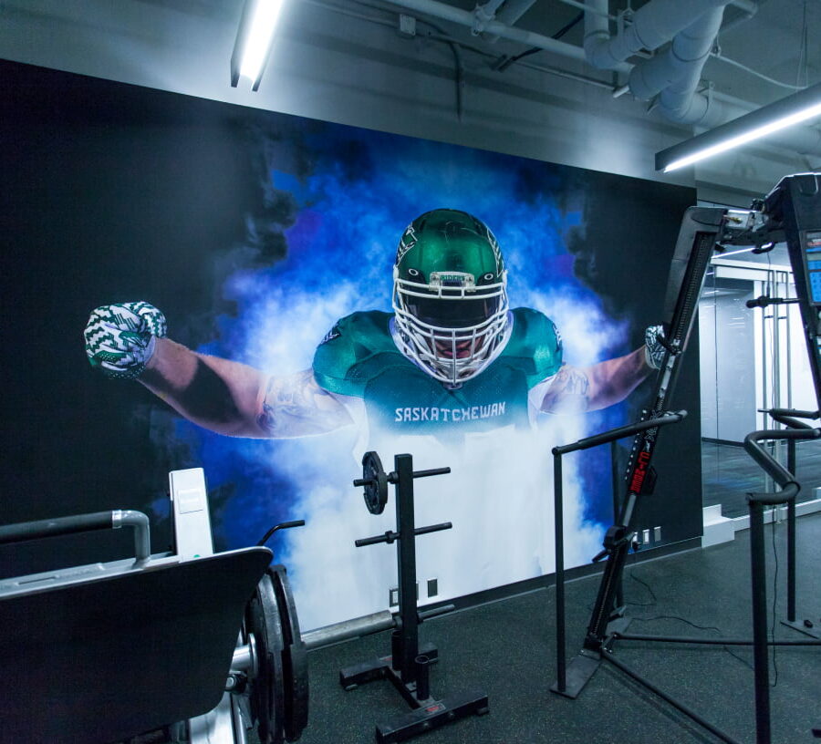 A modern gym features a large wall mural of a football player in a green Saskatchewan uniform emerging from blue smoke, surrounded by various workout machines.