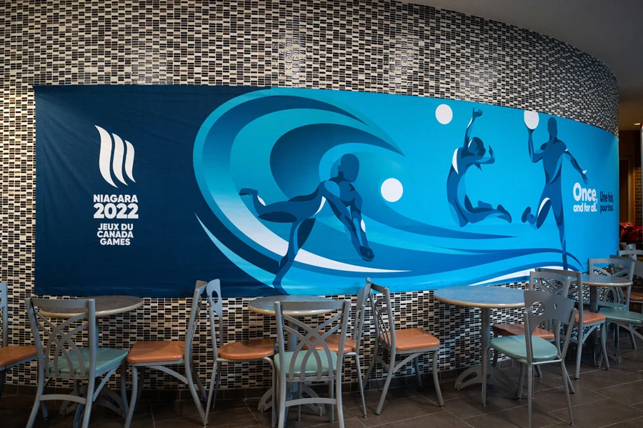 A curved blue mural for the Niagara 2022 Canada Games, depicting abstract athletes and a wave logo, is displayed above tables and chairs in a tiled room.