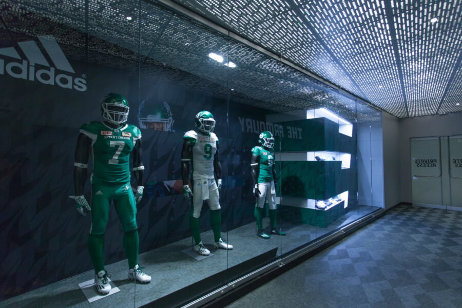 Three mannequins wearing green and white Saskatchewan Roughriders football uniforms are showcased in a glass display case with an Adidas logo on the back wall.