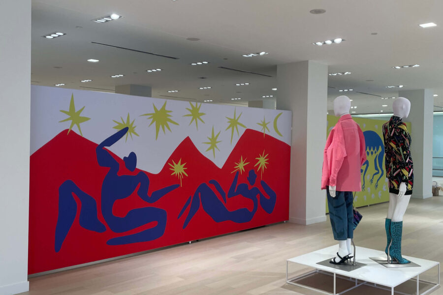 A bright room showcases a large red, blue, and yellow abstract mural with figures and stars, and two mannequins dressed in colorful contemporary fashion on a white platform.