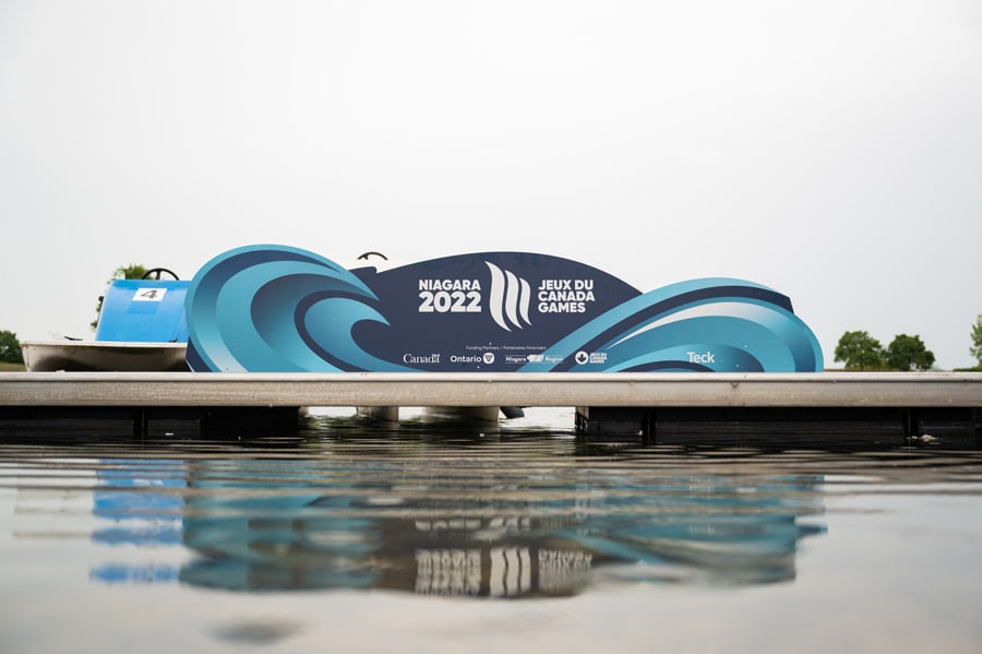 A ""Niagara 2022 Jeux du Canada Games"" banner stands on a dock above reflective water, with a blue paddle boat nearby.