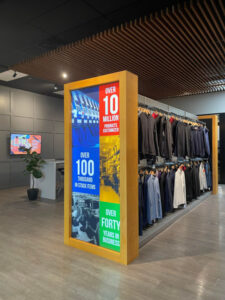 "Modern showroom featuring an illuminated display highlighting company achievements, racks of apparel, and a wooden slatted ceiling."