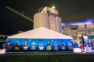 "An outdoor nighttime shot of a tent displaying Canadian Football League players and team logos, with a ""Leon's"" sign on a building and a stadium in the background."