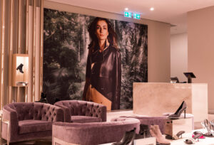 "A luxurious retail interior features purple velvet seating, various shoe displays, and a large wall advertisement of a woman in a leather jacket."