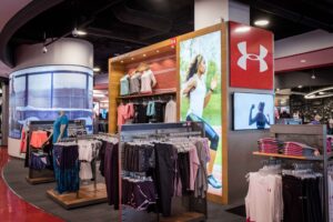 "An Under Armour store section showcasing athletic apparel on racks and mannequins, accompanied by large digital screens displaying brand advertisements."
