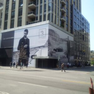 "A building facade prominently displays large black and white ""Holt Renfrew Men"" advertisements with models and a car, above a city street with pedestrians."