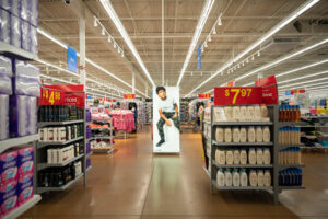 "A brightly lit retail store aisle features shelves of personal care products, clothing racks, and a large advertisement of a boy in the center."