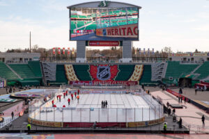 "An outdoor ice hockey rink is being prepared in a stadium for the NHL Heritage Classic 2019, featuring prominent event branding and a large video screen above."
