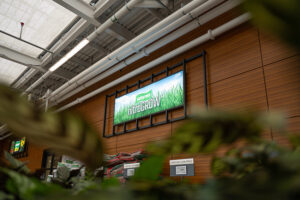 "A digital sign advertising ""Golfgreen nitroGROW"" on a wood-paneled wall, with exposed ceiling pipes and green foliage in the foreground."