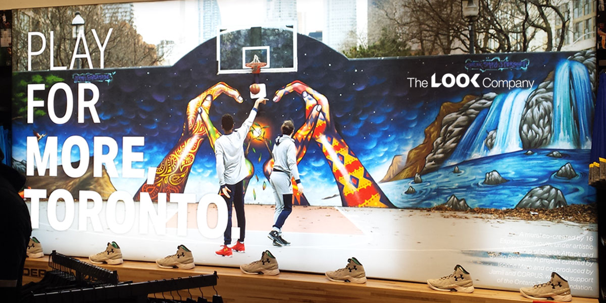 A vibrant mural displays ""PLAY FOR MORE TORONTO"" with two basketball players forming a heart against a starry sky and waterfalls, surrounded by Under Armour shoes on a ledge.