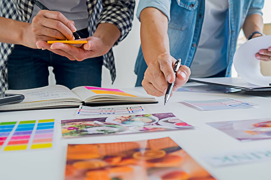 Two individuals collaborate on a design project, reviewing images, color swatches, and notes spread across a table.