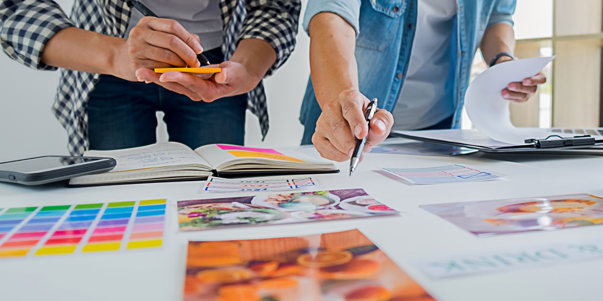 Two individuals collaborate on a design project, reviewing images, color swatches, and notes spread across a table.