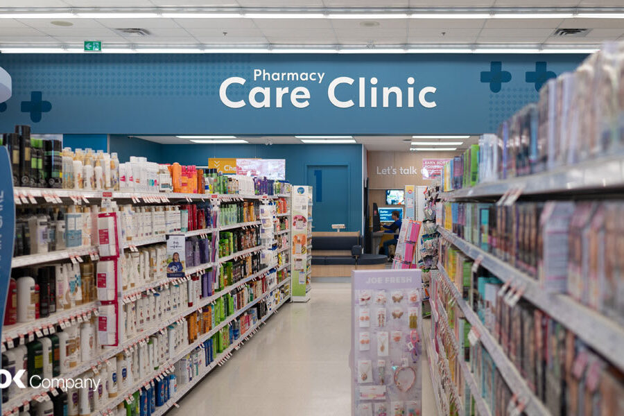 An interior view of a store aisle lined with products, leading to a prominent ""Pharmacy Care Clinic"" sign.